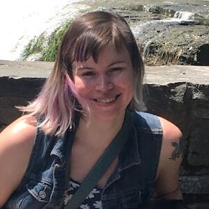 smiling white latine femme in front of a waterfall. their hair is shoulder-length with bangs, brown and pink.